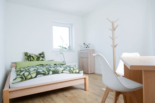 helles, gemütlich eingerichtetes Zimmer in Weiß-, Holz-und Grüntönen mit Bett, Stuhl, Garderobe
